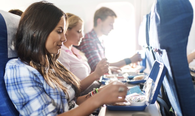 Group of people travelling by airplane. Focus is on foreground, on young woman preparing herself for eating lunch.